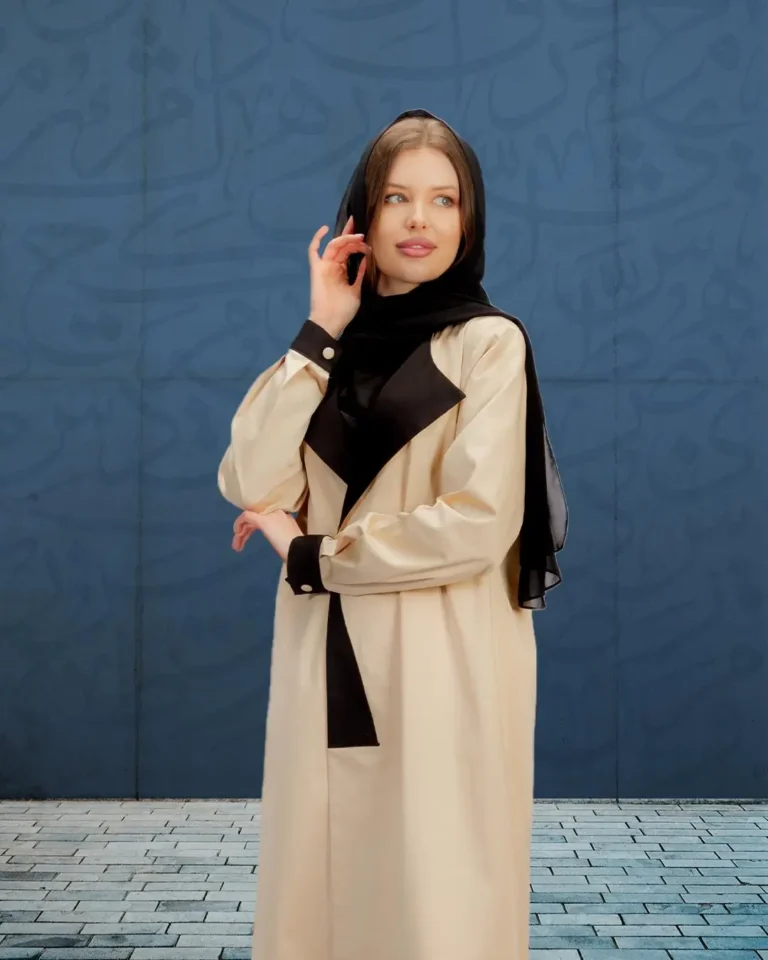 A woman wearing a modern beige abaya with black accents and a black scarf, standing in front of a blue wall with Arabic calligraphy patterns.