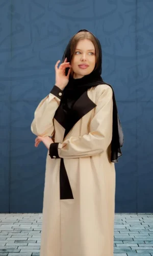 A woman wearing a modern beige abaya with black accents and a black scarf, standing in front of a blue wall with Arabic calligraphy patterns.