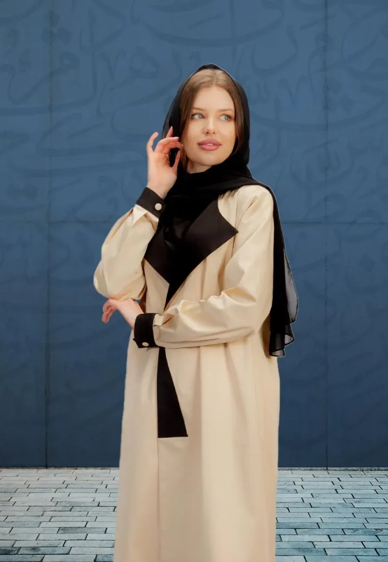A woman wearing a modern beige abaya with black accents and a black scarf, standing in front of a blue wall with Arabic calligraphy patterns.